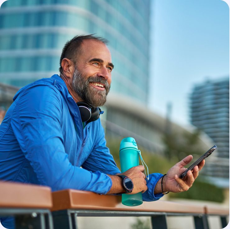 A man in a blue running jacket resting on a railing with headphones, a water bottle, and his phone