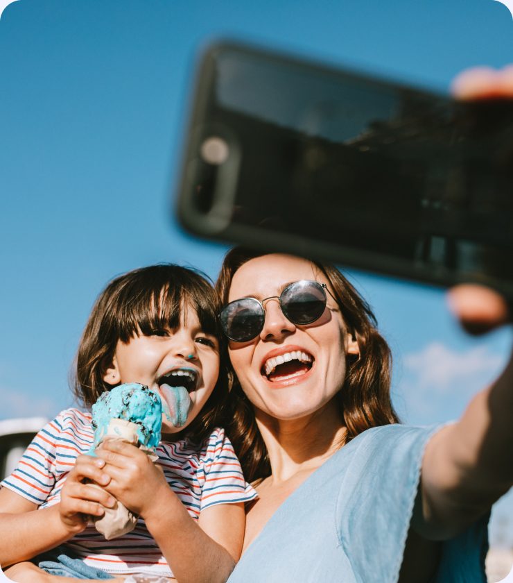 A woman and a child laughing as they take a selfie outdoors, the child eating an ice cream cone