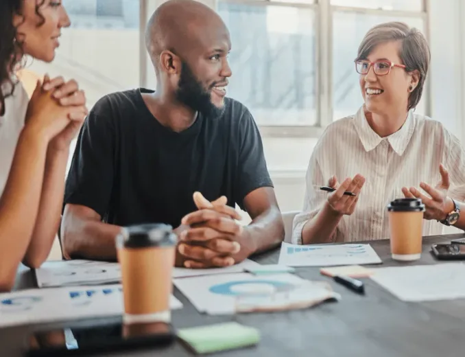 Three colleagues talking and laughing around a table in a bright office
