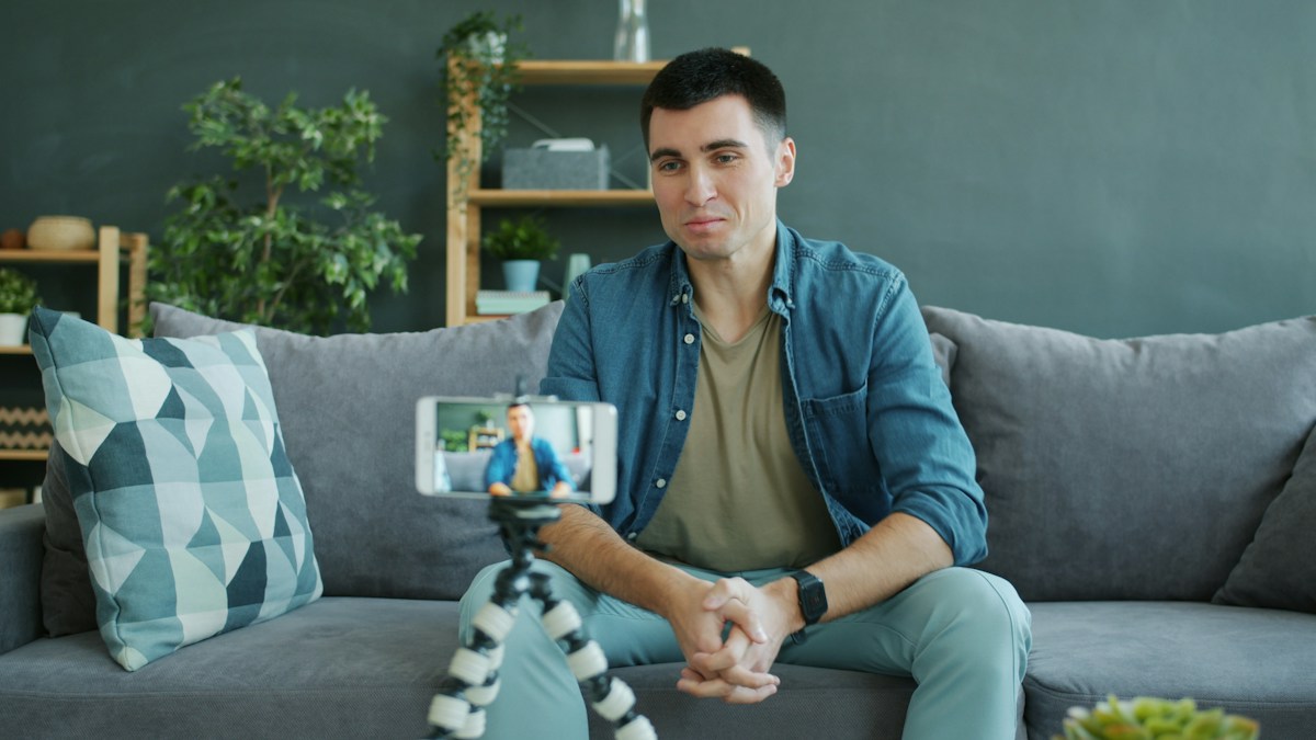 A man on a sofa recording himself talking to a phone mounted on a small tripod.
