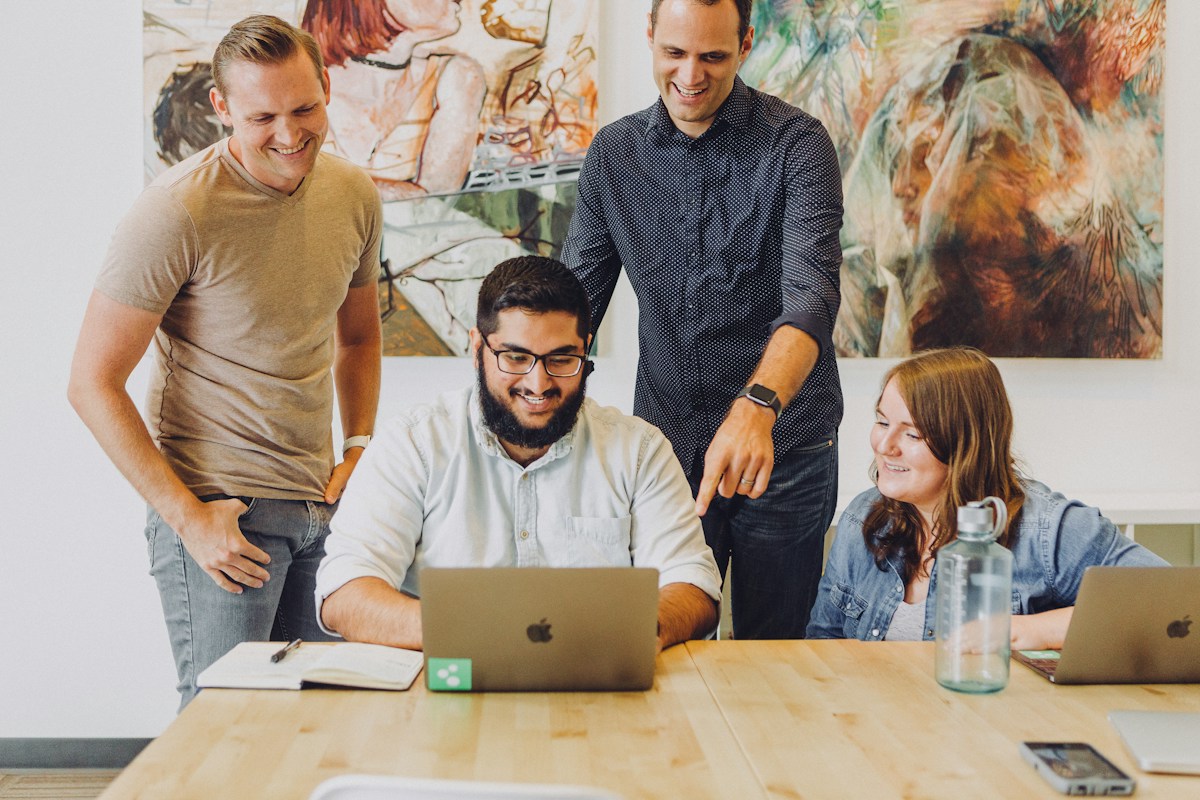 Four colleagues gathered around a laptop at a shared desk, laughing together.