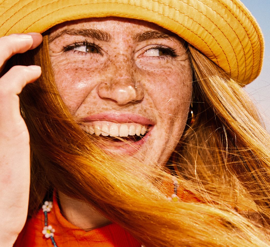 A smiling woman in a yellow sun hat, hair blowing across her face